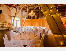 Ein festlich dekorierter Veranstaltungsraum mit großen Holzbalken, einer freiliegenden Backsteinwand und einem Kronleuchter. Runde Tische sind mit weißen Tischdecken, gefalteten Servietten und eleganten Gedecken versehen, während Lichterketten und orangene Vorhänge den Raum schmücken.