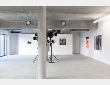 Minimalistisch gestalteter Galerieraum mit Betonboden und einer sichtbaren Betondecke. An den weißen Wänden hängen moderne Kunstwerke, während in der Mitte des Raums eine Skulptur aus metallischen Röhren auf einem Stativ steht. Links sind bodentiefe Glasfenster sichtbar.