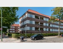 Mehrfamilienhaus mit einer Backsteinfassade und hellen Putzflächen, flachem roten Satteldach und umlaufenden Balkonen mit Blumenkästen. Vor dem Gebäude stehen ein Auto, ein Motorroller sowie Bäume und Hecken entlang eines gepflasterten Gehwegs.