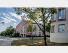 Wohngebiet mit historischen Gebäuden im Stil des frühen 20. Jahrhunderts, darunter ein rosafarbenes Mehrfamilienhaus mit verzierten Fassaden und Erkern. Im Vordergrund stehen Bäume und eine Wiese, flankiert von Gehwegen und einer kleinen Straße.