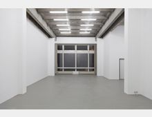 Weißer, minimalistischer Innenraum mit hohen Decken, sichtbaren Betonbalken, grauem Boden und einer großen Fensterfront am Ende des Raumes. Die Beleuchtung besteht aus mehreren schmalen Leuchtstoffröhren an der Decke.