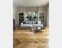 Wohnzimmer mit hellem Parkettboden im Fischgrätmuster, einer großen, beigen Eckcouch vor einem Fenster mit Pflanzen, zwei schwarzen Tischlampen und einem runden Glastisch. Links und rechts vom Fenster befinden sich eingebaute Regalnischen mit Dekoration.