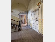 Flur in einem alten Gebäude mit sichtbaren Renovierungsarbeiten. Der Raum zeigt abgenutztes Holzboden, gelb gemusterte Tapeten, eine unvollständig verputzte Türöffnung sowie eine zugemauerte Wand; eine Holztreppe mit dunklem Geländer führt nach oben.