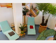 Ruhiger Innenraum mit zwei Liegestühlen, die mit hellblauen Handtüchern bedeckt sind, und einem roten sowie einem grauen Handtuch darauf. Der Raum ist dekoriert mit Topfpflanzen, einer großen Palmenpflanze und einem kreisförmigen Fenster, durch das weitere Pflanzen und Holzelemente sichtbar sind.