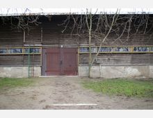 Holzgebäude mit braunen Doppeltüren in der Mitte, umgeben von schmalen Fenstern mit gelben Rahmen im oberen Bereich. Im Vordergrund ist ein mit Gras und Erde bedeckter Hof zu sehen, während kahle Baumzweige den oberen Bildrand einrahmen.