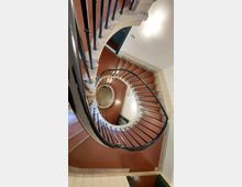 Wendeltreppe in einem mehrstöckigen Gebäude, die sich um eine zentrale Öffnung nach unten windet. Die Treppenstufen sind rotbraun, mit einem Geländer aus schwarzem Metall und hellem Beton.