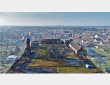 Luftaufnahme eines ländlichen Dorfes mit einer markanten Kirche mit rotem Turm und blauer Kuppel im Vordergrund. Umgeben von mehreren Gebäuden, darunter Wohnhäuser und Scheunen, sowie einer offenen Grünfläche, die von Bäumen gesäumt ist.