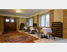 Ein heller Innenraum mit Holzmöbeln, darunter Tische und Stühle mit weißen Tischdecken. Die Wände sind beige, mit großen Fenstern und Weinregalen an den Seiten; ein gemusterter Teppich bedeckt den Boden in der Mitte, und eine hölzerne Doppeltür ist im Hintergrund sichtbar.