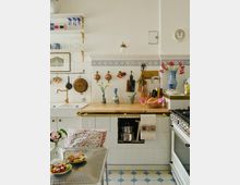 Küche im traditionellen Stil mit weißen Fliesen und blauem Dekor, einer hölzernen Arbeitsplatte, einer weißen Spüle mit goldfarbenen Armaturen und offenen Regalen mit Geschirr und Gläsern. Der Raum enthält dekorative Elemente wie Kupfertöpfe, einen großen blauen Blumenvase mit Blumen und ein Gemälde an der Wand.