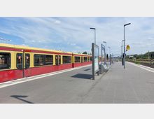 Bahnhofsplattform mit einem gelb-roten S-Bahn-Zug auf der linken Seite. Auf der rechten Seite der Plattform befinden sich Sitzbänke, Fahrplananzeigen und Beleuchtungsmasten. Im Hintergrund sind Gleise und ein Brückenbogen sichtbar, umgeben von vereinzelten Bäumen und Gebäuden.
