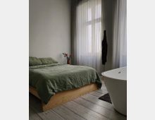 Minimalistisch eingerichtetes Schlafzimmer mit hellem Holzboden, einem hölzernen Bettgestell mit olivgrüner Bettwäsche, einer freistehenden weißen Badewanne und transparenten Vorhängen vor einem großen Fenster. Eine Vase mit Blumen steht auf einem kleinen Tisch neben dem Bett.