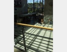 Innenansicht eines modernen Gebäudes mit hoher Glasfassade, durch die Tageslicht einfällt. Im Erdgeschoss sind Sitzbereiche und ein Café oder Kiosk sichtbar, während im Vordergrund ein Geländer und Treppenstufen zu sehen sind.