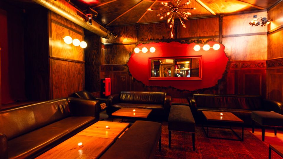 Gemütlicher Bar- oder Lounge-Innenraum mit holzvertäfelten Wänden, Ledersofas und niedrigen Holztischen mit kleinen Kerzen. Warme, gedämpfte Beleuchtung mit Deckenleuchte und Wandlampen, sichtbaren Lüftungsrohren sowie roter Akzentwand mit Spiegel.