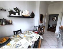 Small kitchen dining area with a rectangular table covered by a patterned tablecloth and black chairs, set against white walls with floating shelves holding plants, jars, and cookware. A tiled floor leads to an open doorway and a narrow sideboard with cups and glassware.