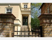 Gelb gestrichene Stadtvilla mit dekorativen Stuckelementen und einem grünen Eingangstor, das über eine Steintreppe erreichbar ist. Im Vordergrund befindet sich ein schmiedeeisernes Tor und eine Backsteinmauer; im Hintergrund sind Bäume sichtbar.