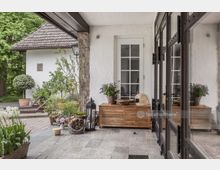 Überdachte Terrasse eines Hauses mit gepflastertem Boden, Holzmöbeln und Dekorationen wie Pflanzen, Körben und Laternen. Im Hintergrund ist ein weißes Gebäude mit Satteldach und umgebendem Garten sichtbar.