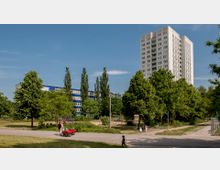 Wohngebiet mit einem hohen Wohngebäude im Hintergrund, umgeben von Bäumen und Grünflächen. Im Vordergrund ein kleiner Spielplatz mit Klettergerüst und spielenden Kindern sowie ein gepflasterter Weg.