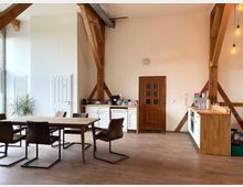 Eine helle Küche mit hohen Decken und sichtbaren Holzbalken. Es gibt eine Küchenzeile mit weißen Schränken, einen Holztisch mit gepolsterten Stühlen sowie eine Holztür in der Wand; rechts hängen dekorative Glühbirnen von der Decke.