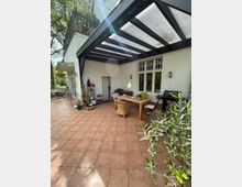 Terrasse mit Terrakottafliesen, überdacht durch ein schwarzes Metallgerüst mit transparenten Kunststoffplatten. Die Terrasse ist mit einem Holztisch, Rattanstühlen, Pflanzen und einem Grill ausgestattet und grenzt an ein weißes Haus mit Sprossenfenstern und einer Tür.