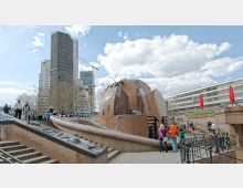 Ein städtischer Platz mit einer großen, kugelförmigen Brunnenanlage aus Metall und Stein, umgeben von Treppen und Sitzbereichen. Im Hintergrund sind hohe, moderne Bürogebäude sowie ein Kran zu sehen, und Menschen spazieren oder sitzen in der Umgebung.