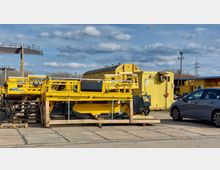 Ein Industriestandort mit gelben, auf Paletten gelagerten Hebe- und Wartungsplattformen mit der Aufschrift „Palfinger Railway“, einem gelben Container im Hintergrund und einem geparkten blauen Auto rechts im Bild. Der Himmel ist leicht bewölkt, und im Hintergrund sind Bäume sowie Masten sichtbar.