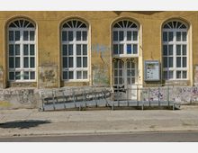 Gelb gestrichene Fassade eines alten Gebäudes mit hohen, weißen Sprossenfenstern und einer Tür in der Mitte, die von Graffiti bedeckt ist. Vor dem Gebäude befindet sich eine Rampe mit einem Metallgeländer; ein Schaukasten mit der Aufschrift „Galerie im Prater“ hängt rechts neben der Tür.