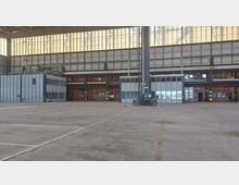 Große, leere Industriehalle mit hohen Decken, breiten Glas- und Metallkonstruktionen sowie einer Reihe orangefarbener Türen und Fenster auf der Rückwand. Im Vordergrund ist ein weitläufiger Betonboden mit eingeritzten Linien und wenigen Gegenständen sichtbar.