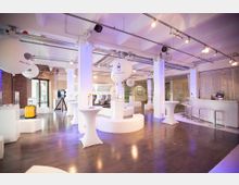 Moderner Veranstaltungsraum mit hellen Wänden, Holzboden und weißen Stehtischen. Der Raum ist mit dekorativen weißen Säulen, Deckenbeleuchtung und einigen technischen Exponaten ausgestattet, darunter Sitzmodelle und ein gelber Koffer.