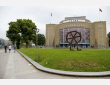 Außenansicht der Volksbühne in Berlin, einem historischen Theatergebäude mit einer Fassade aus Sandstein und einem großen Metallskulptur-Rad auf einer Rasenfläche davor. Im Vordergrund führt ein Gehweg entlang, umgeben von Grünflächen und Bäumen.
