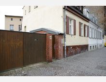 Ein Wohngebäude mit hell beiger Fassade, Fenstern mit braunen Klappläden und einer Wand aus Ziegelsteinen im unteren Bereich; daneben ein hoher hölzerner Torabschluss und ein kleiner Ziegelanbau mit Hausnummer "34". Im Vordergrund ist ein gepflasterter Gehweg sichtbar.