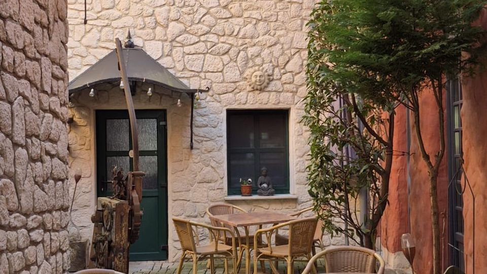Kleiner Innenhof mit Kopfsteinpflaster und mehreren runden Café-Tischen mit Korbstühlen, umgeben von Naturstein- und rot verputzten Wänden. Im Hintergrund stehen eine grüne Eingangstür mit kleinem Vordach und Fenster; an der rechten Wand wachsen Kletterpflanzen und ein kleiner Baum.