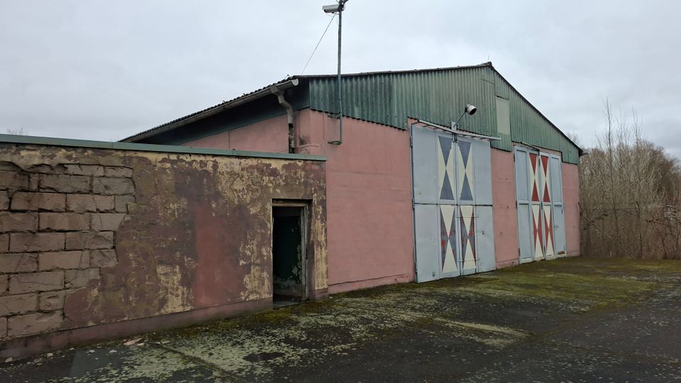 Außenansicht einer einfachen Lager- oder Industriehalle mit grünem Wellblechdach und rosafarbenen Wänden, davor ein kleiner Anbau mit verwitterter Fassade und offener Tür. Große Metall-Schiebetore mit geometrischen X-Mustern und ein Lampenmast stehen vor einem asphaltierten Hof, im Hintergrund sind kahle Bäume und ein grauer Himmel zu sehen.
