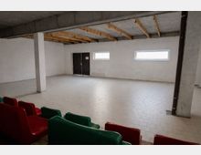 Großer, leerer Innenraum mit gefliestem Boden, weiß verputzten Wänden und Beton-Decke mit sichtbaren Holzbalken. An der Rückwand befinden sich eine schwarze Doppeltür und zwei schmale, horizontale Fenster; im Vordergrund sind Reihen von roten und grünen Kunststoffsitzen zu sehen.