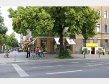 Straßenszene mit einem Eckgebäude, auf dem das Schild „Kreuzberg Apotheke“ zu sehen ist, umgeben von Bäumen. Vor dem Gebäude befinden sich Gehwege, Fahrräder und Fußgänger, im Hintergrund sind Cafés mit Außenbestuhlung erkennbar.