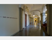 Breiter, glänzender Flur eines historischen Gebäudes mit hohen gewölbten Decken, Säulen und hölzernen Türen; Wandschilder und Beschriftungen weisen auf Raumnummern hin, während Hängelampen den Raum beleuchten. Im Hintergrund sind eine Treppe und weitere Türen sichtbar.