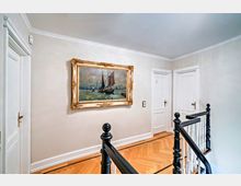 Flur mit Holzboden in Fischgrätmuster, weiß gestrichenen Wänden und schwarzen Holzgeländern. An einer Wand hängt ein gerahmtes Gemälde mit einer maritimen Szene, daneben befinden sich zwei weiße Türen mit goldenen Türgriffen.