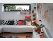 Wohnzimmer mit hellem, modernem Design; eine weiße Eckcouch mit gemusterten und farbigen Kissen steht vor einem breiten Fenster mit Pflanzen auf der Fensterbank, daneben weiße Regale mit Büchern und Dekorationen. An der Wand hängt eine abstrakte Kunsttapete, und auf einem weißen Beistelltisch stehen rote Blumen in einer Vase.