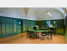 Ein Büroraum mit grüner Wandverkleidung und einem Gewölbedecken-Design. In der Mitte stehen ein grüner, winkelförmiger Schreibtisch und mehrere schwarze Stühle; an den Wänden befinden sich Garderobenhaken, eine Tür und eine Büroecke mit Lampe, Dokumenten und Monitor.