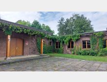 Backsteinbau mit efeubewachsenen Fassaden, Holzfenstern und einer großen Holztür. Der Hof ist mit Pflastersteinen ausgelegt, und im Hintergrund sind Bäume und ein bewölkter Himmel zu sehen.