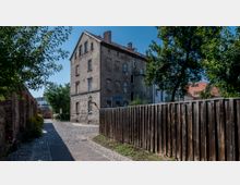 Mehrstöckiges Wohnhaus aus Backstein an einer gepflasterten Straße, umgeben von Bäumen, einem Holzzaun und angrenzenden gemauerten Gebäuden; im Hintergrund sind weitere Häuser mit roten Dächern sichtbar.