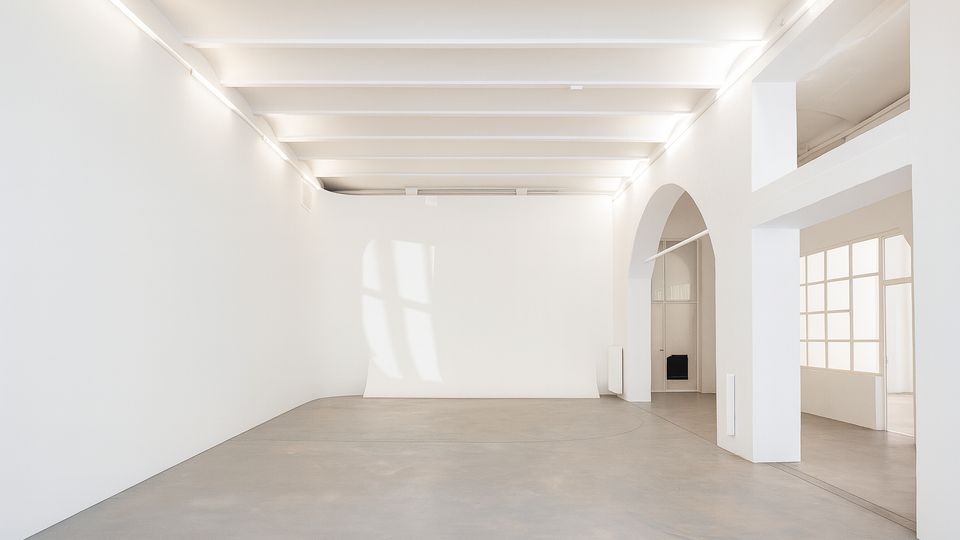 Heller, minimalistischer Innenraum mit weißen Wänden, sichtbaren Deckenbalken und glattem Betonboden. Rechts führen ein großer Rundbogen und verglaste Tür- und Fensterflächen in angrenzende Räume.