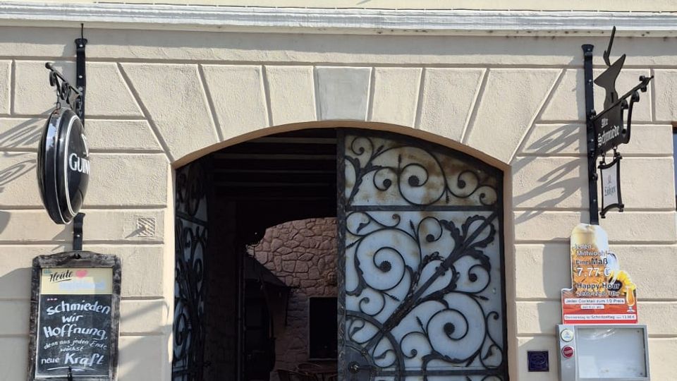 Eingangsfassade eines hellen, zweigeschossigen Gebäudes mit dem Schriftzug „Alte Schmiede“ über einem großen Rundbogenportal. Das Portal hat ein geöffnetes, schmiedeeisernes Gittertor mit Ornamenten und führt in einen gepflasterten Durchgang; seitlich hängen Schilder und ein Aushangkasten.