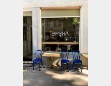 Außenansicht eines Cafés mit großer Glasscheibe und dem Schriftzug "SFERA" auf der Fensterscheibe. Vor dem Café stehen drei blaue Stühle und zwei kleine gelbe Wandtische, die auf einem gepflasterten Gehweg platziert sind.