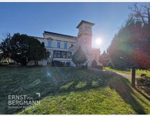 Historisches Gebäude mit hellem Putz und einer markanten Turmstruktur, umgeben von gepflegtem Rasen und hohen Bäumen; im Vordergrund ein Weg, Sonnenstrahlen und ein klarer blauer Himmel im Hintergrund. Text „Ernst von Bergmann Klinikum Potsdam“ ist am unteren linken Bildrand sichtbar.