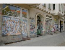 Fassade eines älteren Gebäudes in einer urbanen Umgebung, bedeckt mit Graffitis, Plakaten und Abnutzungsspuren. Die Fenster, Türen und Wände zeigen verschiedene Schichten von Farben und beklebten Papierresten.
