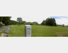 Flache Wiesenlandschaft mit einzelnen Bäumen am Horizont, flankiert von einem asphaltierten Weg links und einer Reihe großer Steine. Im Vordergrund steht ein grauer Gedenkstein mit Aufschrift und farbigem Logo der Region "Lebuser Land".
