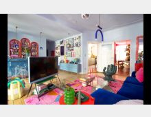 Wohnzimmer mit farbenfroher Einrichtung, bestehend aus einem blauen Sofa, einem pinken Teppich, einer Glas-Couchtischplatte, einem TV auf einem schmalen Tisch und verschiedenen modernen Dekorationen. Im Hintergrund sind eine kleine Bar mit leuchtend rotem Rahmen, eine Kommode mit Wanddekorationen und ein Durchgang zur beleuchteten Küche zu sehen.