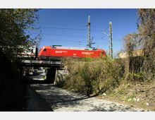 Eine rote Lokomotive fährt auf einer Eisenbahnbrücke über eine gepflasterte Unterführung. Im Vordergrund sind überwucherte Sträucher und Laubbäume, während im Hintergrund Strommasten und blauer Himmel sichtbar sind.