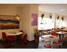 Ein gemütliches Café mit einer Mischung aus Retro-Möbeln, darunter rote Stühle an einem weißen runden Tisch und ein weiterer Tisch mit weißem Metallstuhl. Im Hintergrund sind große Fenster mit transparenten Vorhängen, dekorativen Pflanzen und weiteren Sitzgelegenheiten zu sehen.