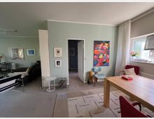 Heller Wohn- und Essbereich mit hellgrünen Wänden, großem Fenster mit Vorhang und Teppichboden; an den Wänden hängen gerahmte Bilder und ein großes farbiges Gemälde. Rechts steht ein Holztisch mit roten Stühlen, daneben ein Sitzbereich mit Sofa, Beistelltisch und weiteren Stühlen sowie eine offene Tür zum Flur.